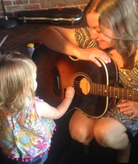 Linda Bassick holds her acoustic guitar while a toddler strums the strings.