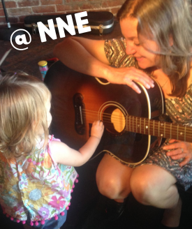 Photo of Linda with a guitar and a toddler reaching out to strum the strings